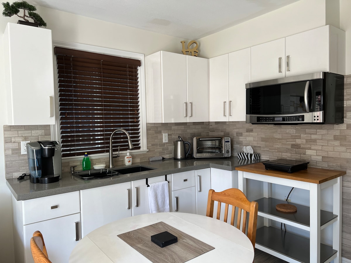 The well-equipped kitchen features modern white cabinetry and a countertop with a neutral stone backsplash. A wooden dining table with chairs is situated nearby. Essential appliances, including a microwave and coffee maker, are visible, along with natural light filtering through the window with wooden blinds.
