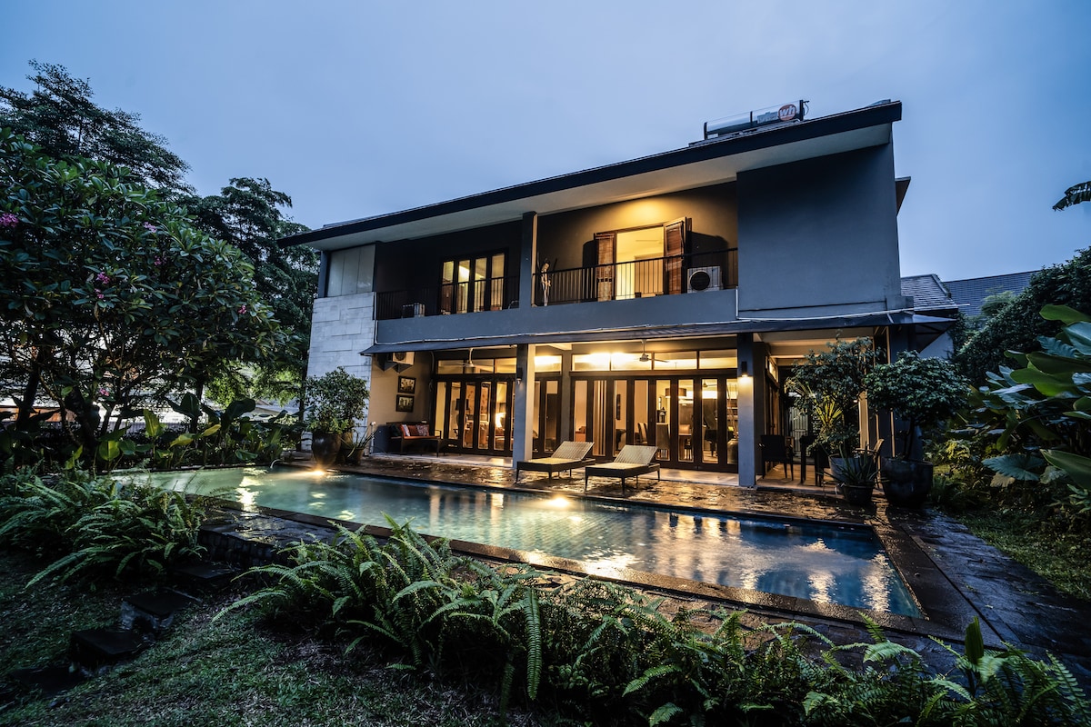 The architectural design of the house is showcased in the evening, with warm lights illuminating the expansive balconies. A serene swimming pool is bordered by lush greenery, enhancing the tranquil outdoor space. The structure features large glass doors, integrating the indoor and outdoor areas.