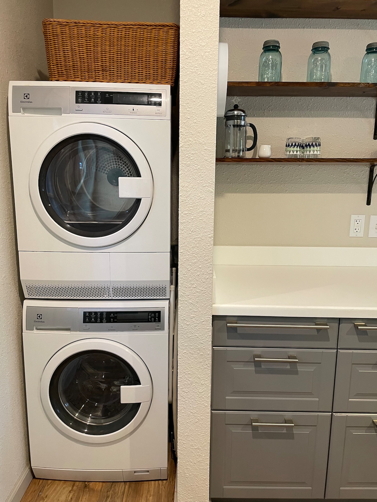 The laundry area features a stacked washer and dryer in a compact space, accompanied by shelves holding glass canisters. A clean countertop provides additional surface area, while gray cabinetry offers storage options beneath.