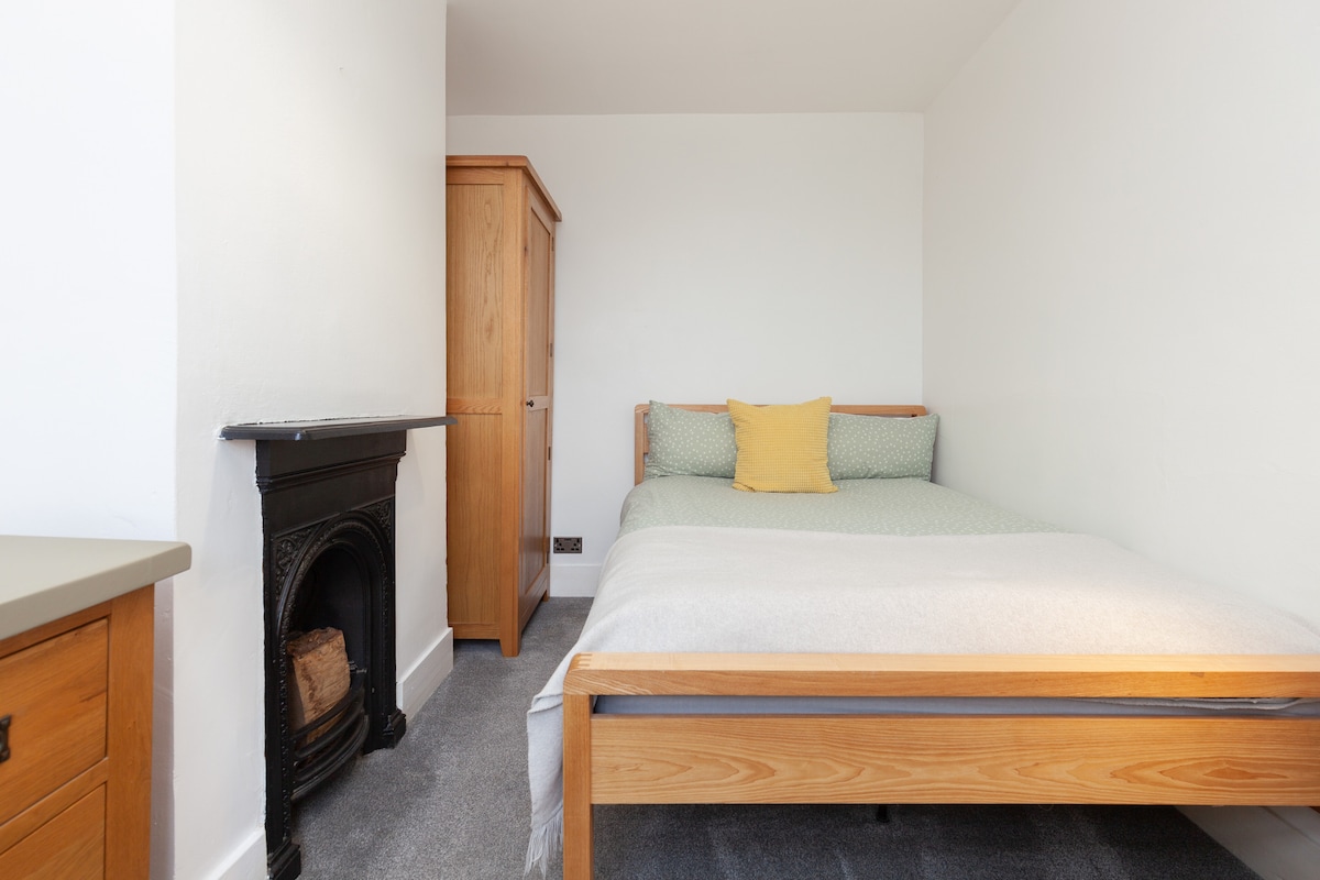 A spacious bedroom features a wooden bed with a light green bedspread and a yellow accent pillow. A wooden wardrobe stands against the wall, and a decorative fireplace adds character. Soft gray carpeting complements the neutral walls, creating a calming environment.