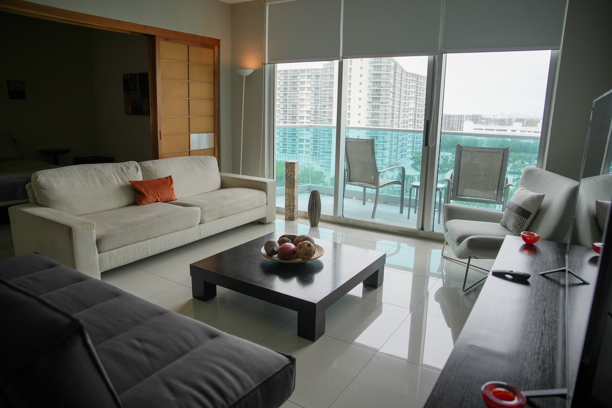 A spacious living area is furnished with a light-colored sofa and a dark coffee table, complemented by decorative items. Large windows allow natural light to fill the room, revealing views of the balcony and surrounding area. Stylish decor is evident throughout.