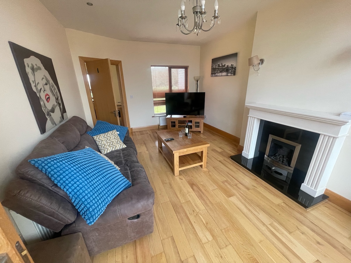 The living area features a comfortable sectional sofa adorned with a blue throw and decorative pillows. A wooden coffee table is placed in front, accompanied by a flat-screen television. Natural light enters through a window, and a simple fireplace enhances the ambiance.