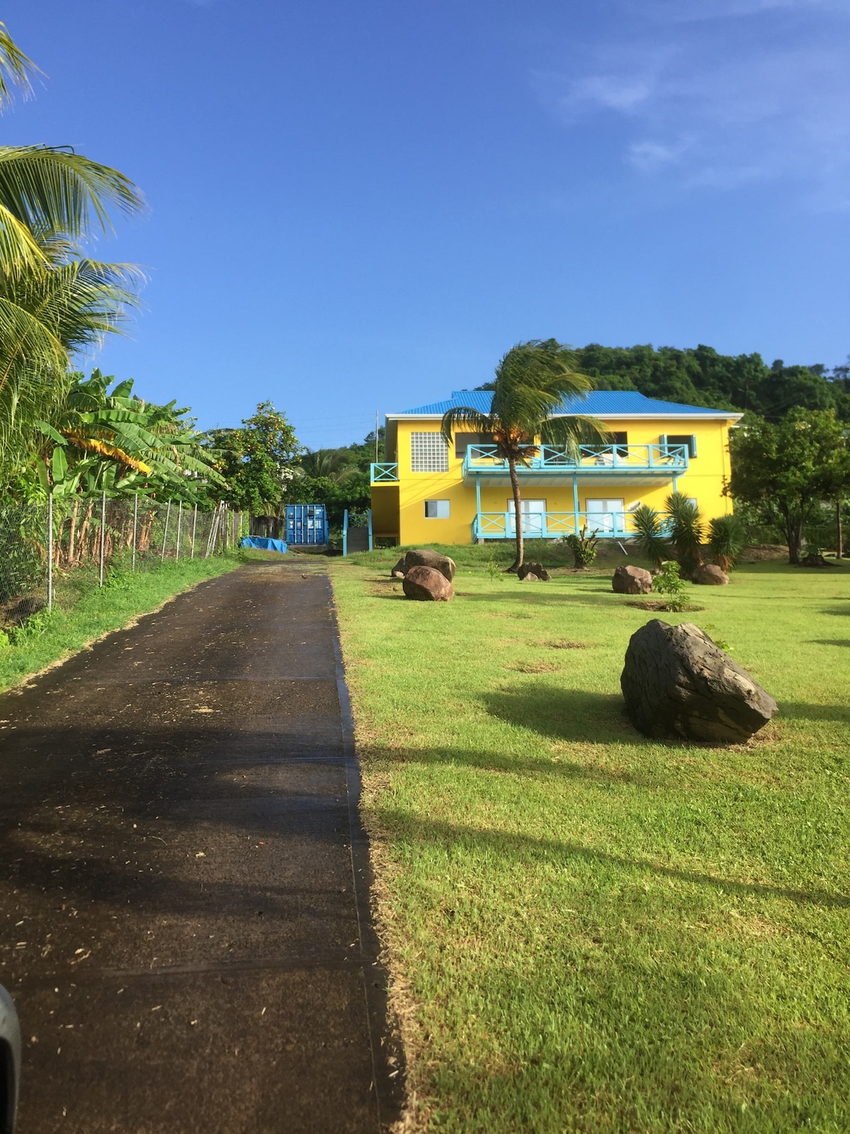 A vibrant yellow building is captured against a clear blue sky, surrounded by lush greenery. A paved pathway leads towards the entrance, flanked by several large rocks on well-maintained grass. Palm trees sway gently in the breeze, contributing to a serene atmosphere.