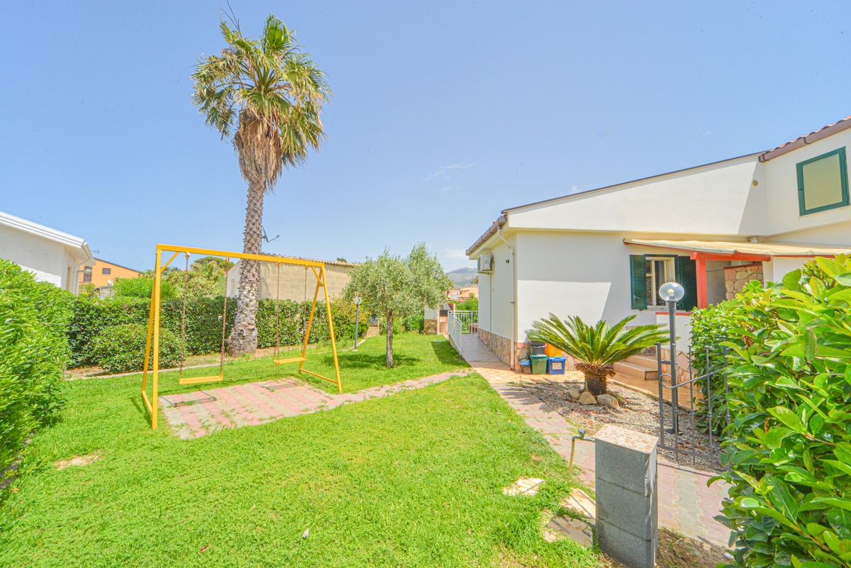 A well-maintained garden is visible, featuring a yellow swing set surrounded by green grass. Lush shrubs and a palm tree add to the scenery, while the villa can be seen in the background with its welcoming entrance.