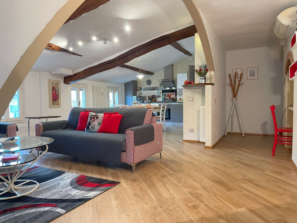 A spacious living area features wooden beams overhead and a comfortable sofa adorned with decorative cushions. A glass coffee table rests atop a patterned rug. The open layout provides a clear view of the modern kitchen in the background, enhancing the bright and airy atmosphere.