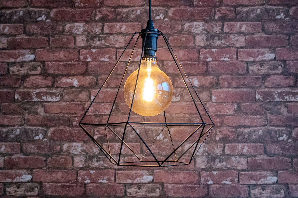 A geometric pendant light hangs from the ceiling, featuring a warm glowing bulb encased in a black wire frame. The light contrasts against a backdrop of textured red brick wall, adding visual interest to the space.