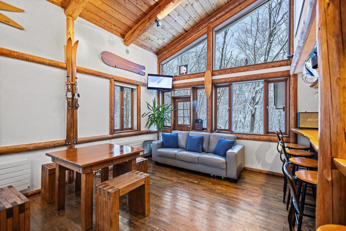 The spacious lounge area features large windows that provide views of the snow-covered outdoors. A comfortable sofa is positioned near a wooden dining table, complemented by bench seating. The room includes wooden beams and a ski-themed decor, creating a cozy atmosphere.