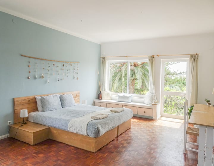 Ocean Suite's Master bedroom, with a king-sized bed, large windows overlooking the garden & sea, and a built-in window nook. French doors lead to a private balconette shared with Bedroom 2. Bright, airy, and beachy with dainty decorative touches.