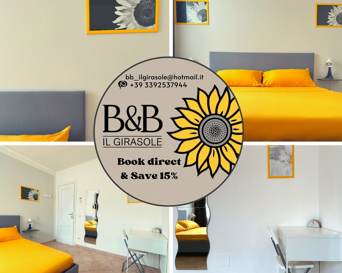 A well-lit bedroom features a spacious bed with bright yellow bedding. A minimalist desk and chair are positioned beside the bed. The walls are adorned with framed artwork, while a mirror reflects the room's inviting atmosphere.