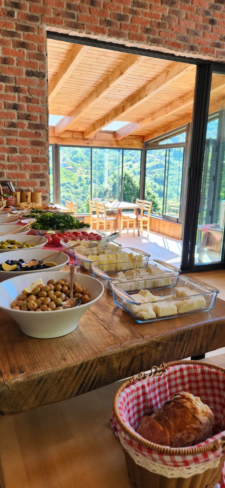 A wooden table displays a variety of food items, including olives, cheese, and fresh bread. Large glass doors provide a view of the surrounding landscape, while the space maintains a bright and airy feel with natural light streaming in.