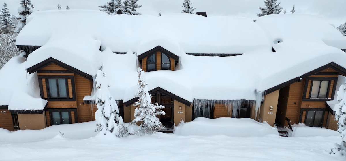 A snow-covered exterior of a condo complex is presented, with roofs blanketed in white snow and icicles hanging from the eaves. Trees dusted with snow surround the building, creating a serene winter scene.