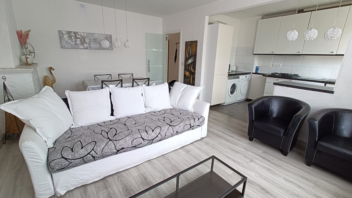 The living area features a white sofa adorned with decorative pillows and a patterned throw. Two dark chairs are positioned nearby, alongside a coffee table. A dining area with metal chairs is visible in the background, complemented by white cabinetry and a washing machine.