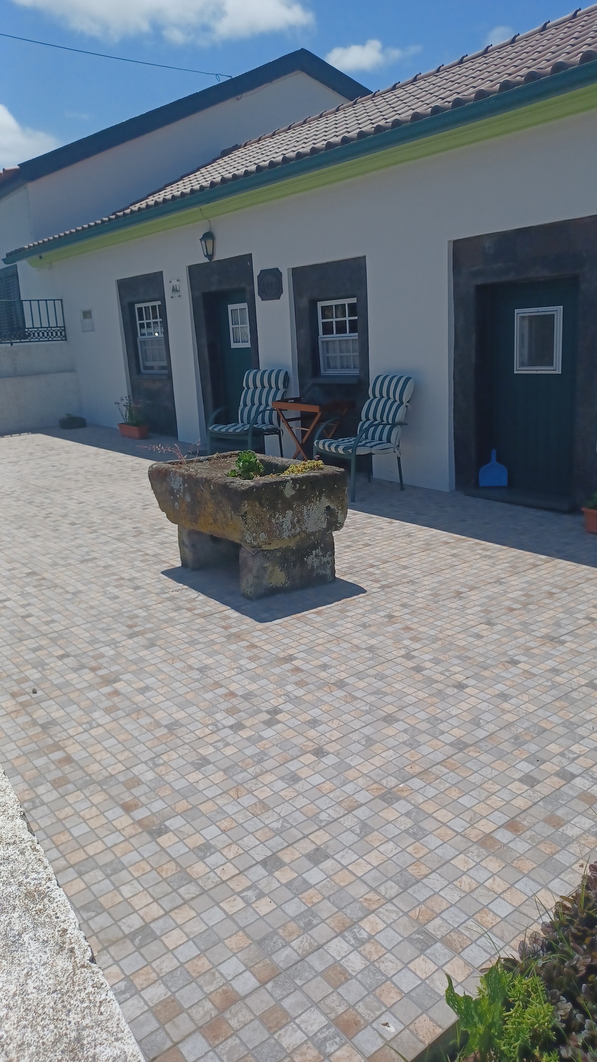 A tiled courtyard features a rustic stone feature at its center, complemented by two chairs arranged against the wall. Doorways leading into the house are visible, framed by greenery and clear skies above, creating a welcoming outdoor space.
