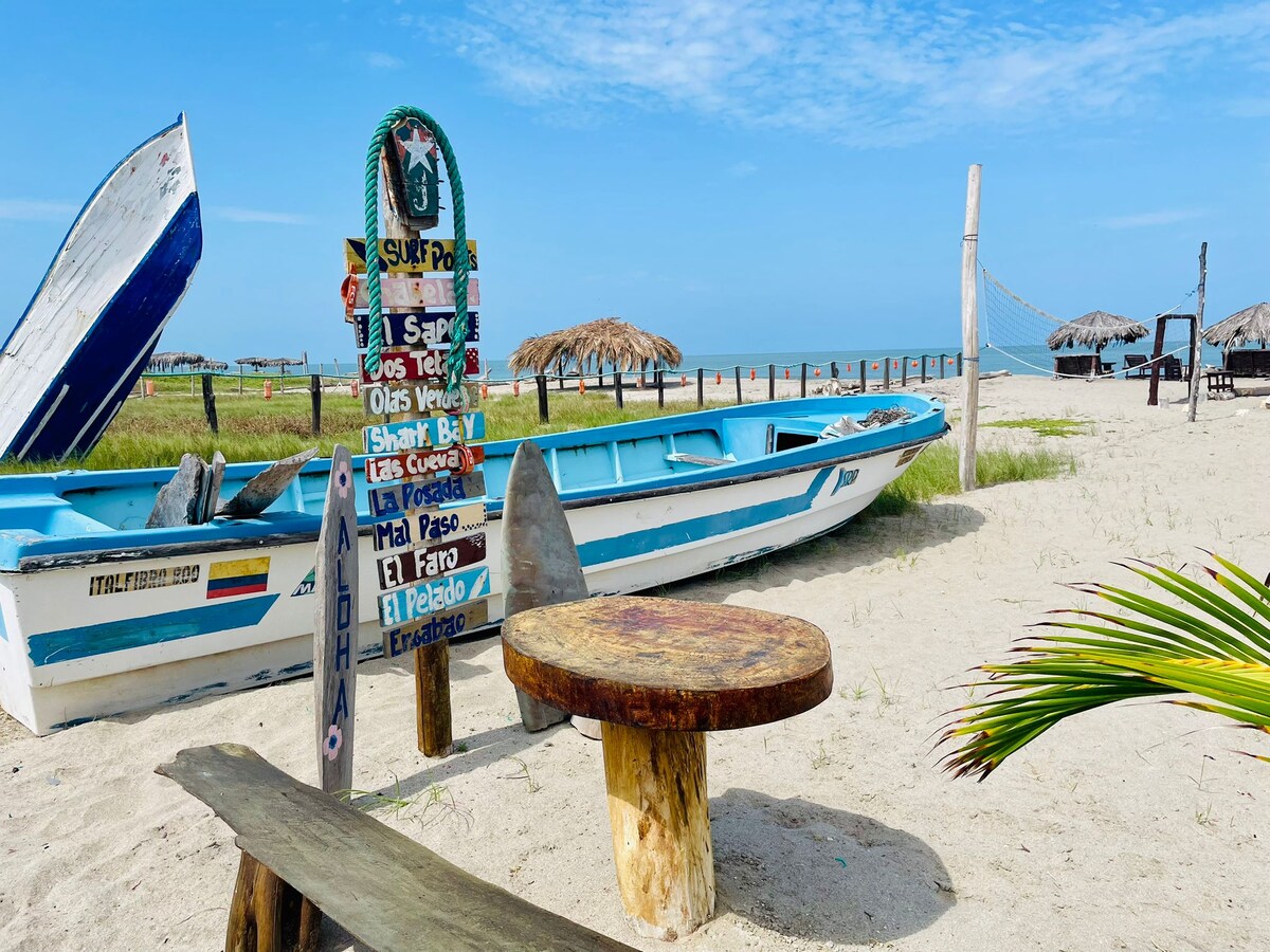 A rustic wooden table sits on the sandy beach, accompanied by two fishing boats nearby. A sign with various beach destinations stands prominently, with palm-thatched huts in the background. Along the horizon, the ocean meets a clear blue sky.
