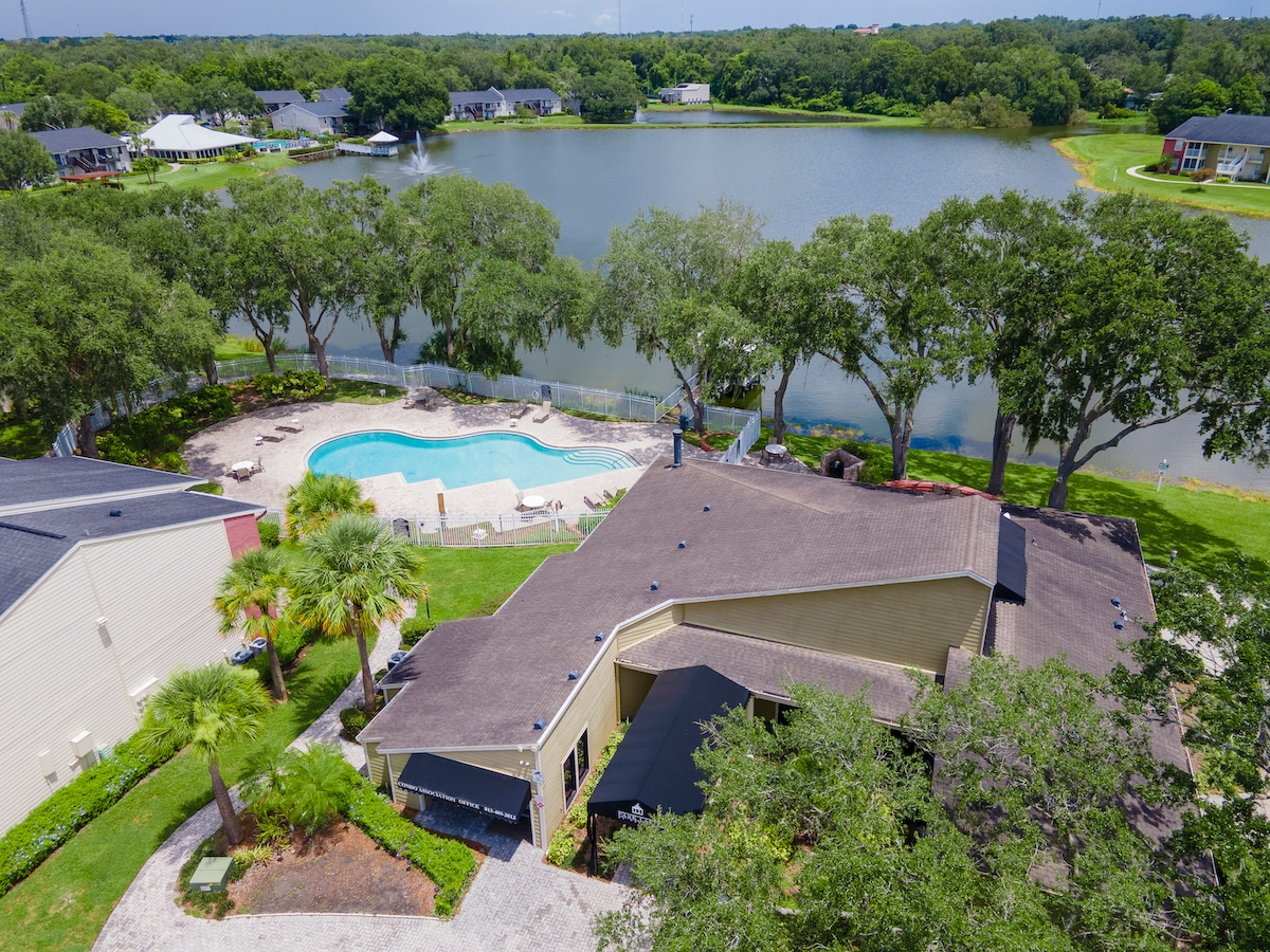 An aerial view displays a beautiful pool area surrounded by lounge chairs and palm trees, adjacent to a calm water body. The landscape showcases green trees and grassy areas, providing a tranquil environment. Nearby buildings complement the serene setting.