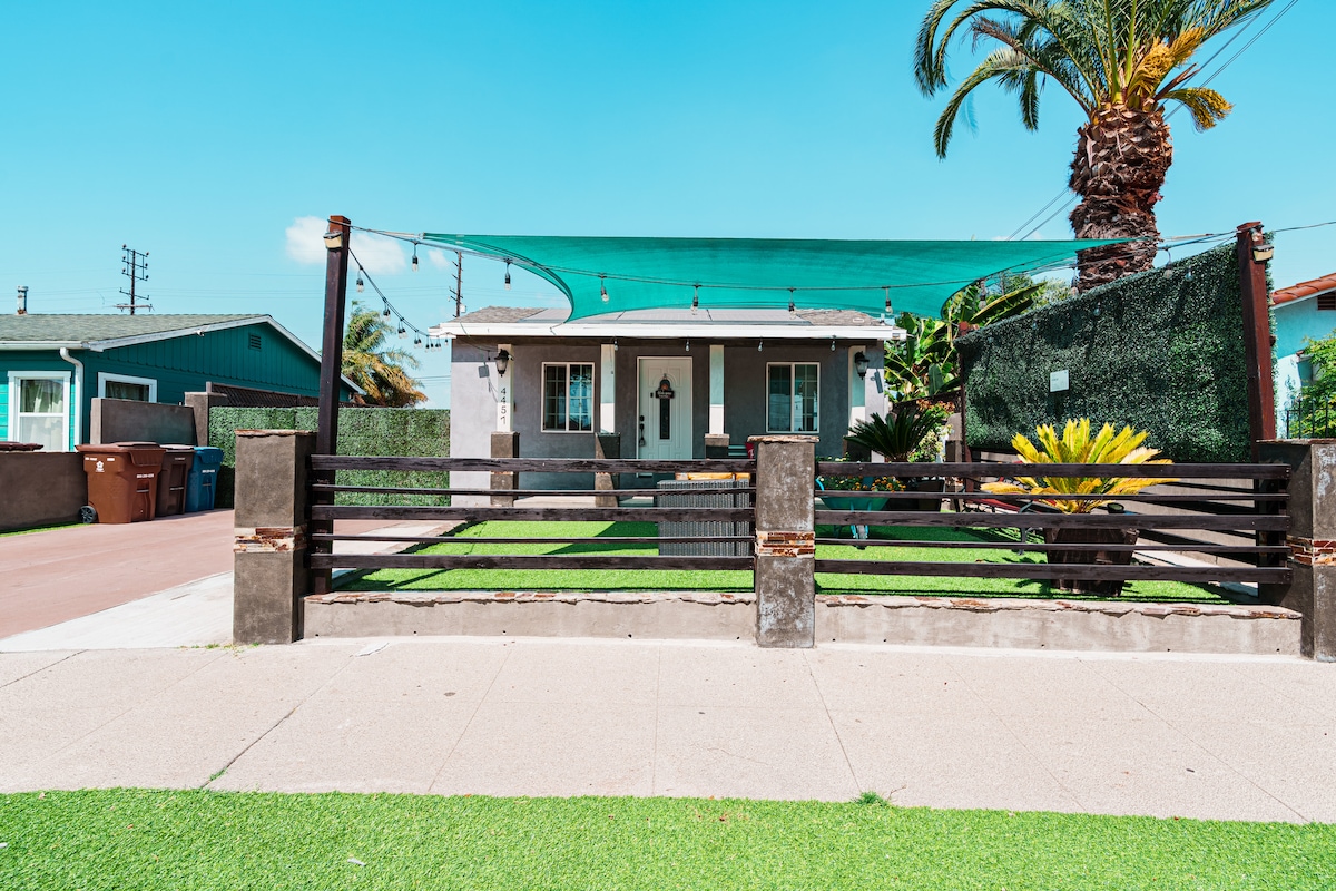 The house is set back from the street, framed by tall palm trees and surrounded by lush green landscaping. A shaded patio area extends over the entrance, providing a welcoming outdoor space. The house features a simple, modern facade with a distinct arrow design above the door.