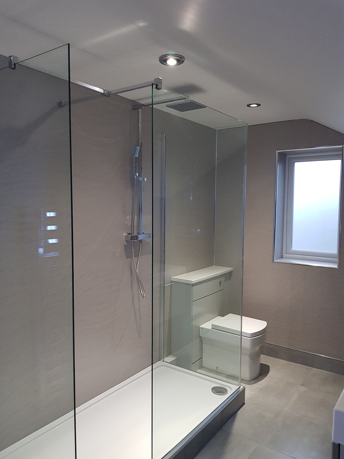 A modern wet-room style shower is enclosed by clear glass panels, showcasing a sleek design. A minimalistic toilet is positioned beside custom cabinetry. Natural light enters through a small window, enhancing the room's bright and clean appearance.