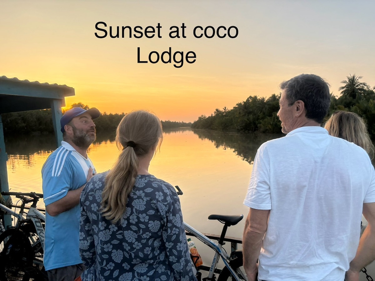 A serene sunset reflects on calm waters, creating a warm golden glow. Silhouetted figures stand along a dock, surrounded by lush greenery. Bicycles are parked nearby, indicating an inviting outdoor space for leisure and relaxation.