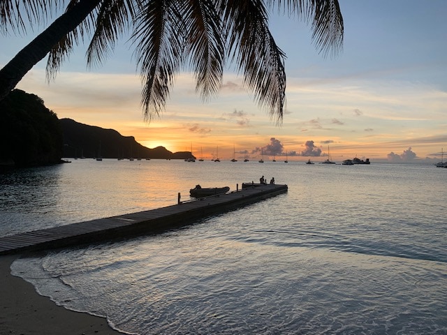 A serene sunset view features a wooden dock extending into calm waters, with silhouettes of distant boats anchored in the bay. Gentle waves reflect the soft hues of the sky, while a palm tree frames the scene, enhancing the peaceful coastal atmosphere.