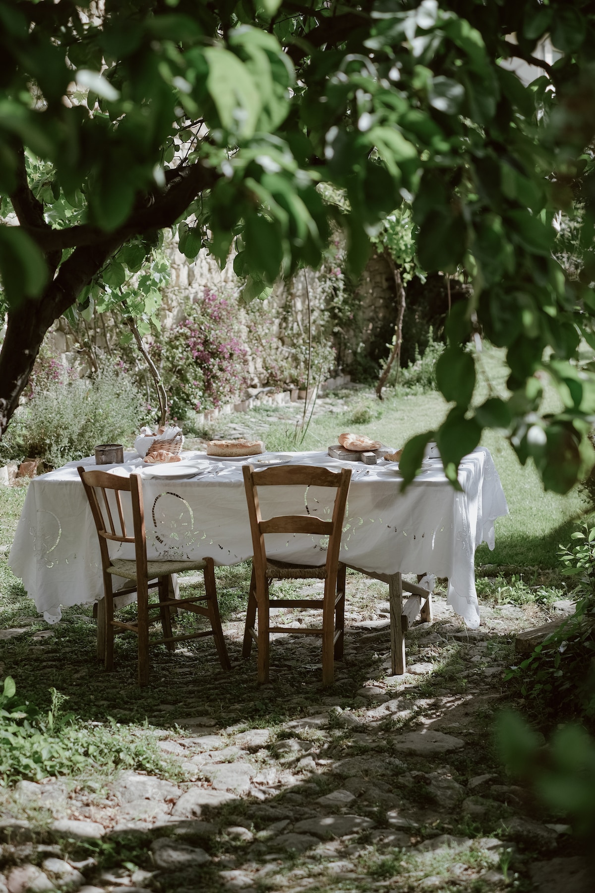 A wooden table is set under a tree, surrounded by four chairs. A white tablecloth covers the table, which is adorned with simple plates and bread. The scene is framed by lush greenery, creating a tranquil setting for outdoor dining.