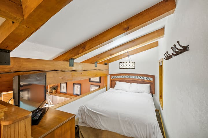 Спална соба на поткровје со удобен брачен кревет (широк 150-179 см) и телевизор, сместена над дневниот простор за приватно, но поврзано место за одмор.