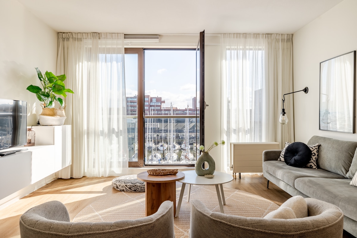 A comfortable living area is highlighted by large windows that open to a harbor view. A grey sofa and coordinating lounge chair provide seating, while a round coffee table and decorative accents enhance the space. Natural light fills the room, complemented by airy curtains.