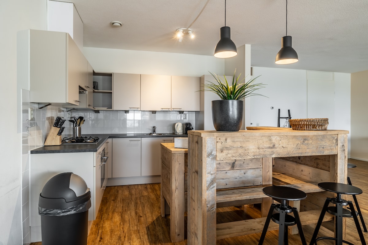 A spacious kitchen area features modern appliances and light cabinetry. A large wooden dining table made from reclaimed wood is surrounded by black stools, creating a casual dining atmosphere. Natural light is allowed in through a nearby window, enhancing the functional layout.