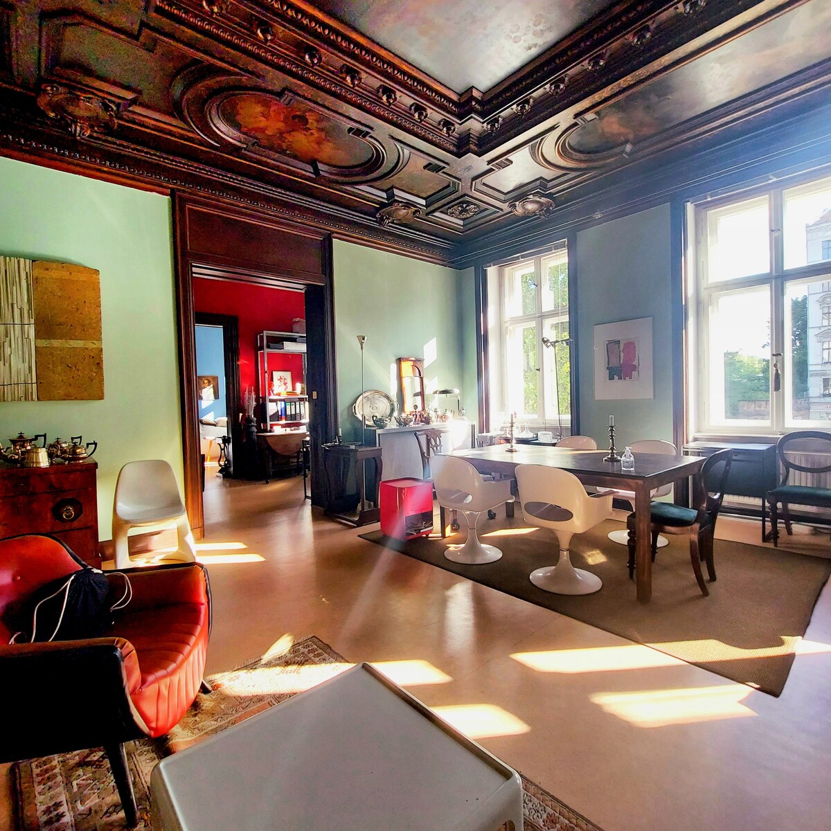 A spacious living area features high ceilings adorned with intricate ceiling designs. Natural light streams through large windows, illuminating the eclectic mix of furniture, including modern and traditional seating. A dining table is situated centrally, complemented by soft, neutral tones on the walls.