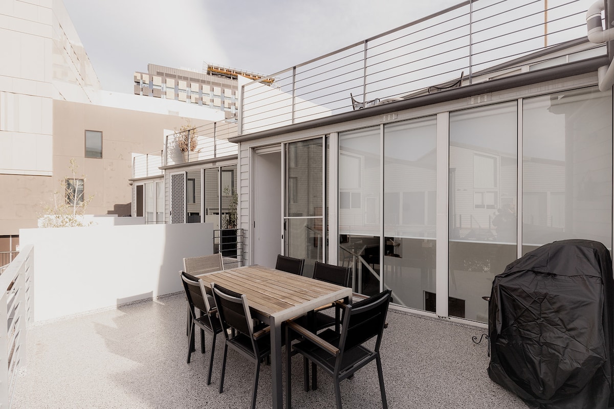 An outdoor terrace features a large wooden dining table surrounded by black chairs, offering ample space for dining. A BBQ grill stands on one side, with the area surrounded by low walls for privacy. Glass sliding doors provide access to the indoor space.