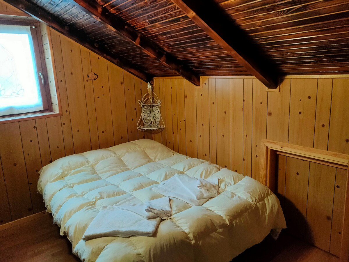 A cozy bedroom is presented with wooden paneling and a sloped ceiling. A spacious bed covered with a quilted comforter is centered in the room. Natural light enters through a small window adorned with a lace curtain, while a decorative pendant light hangs above.