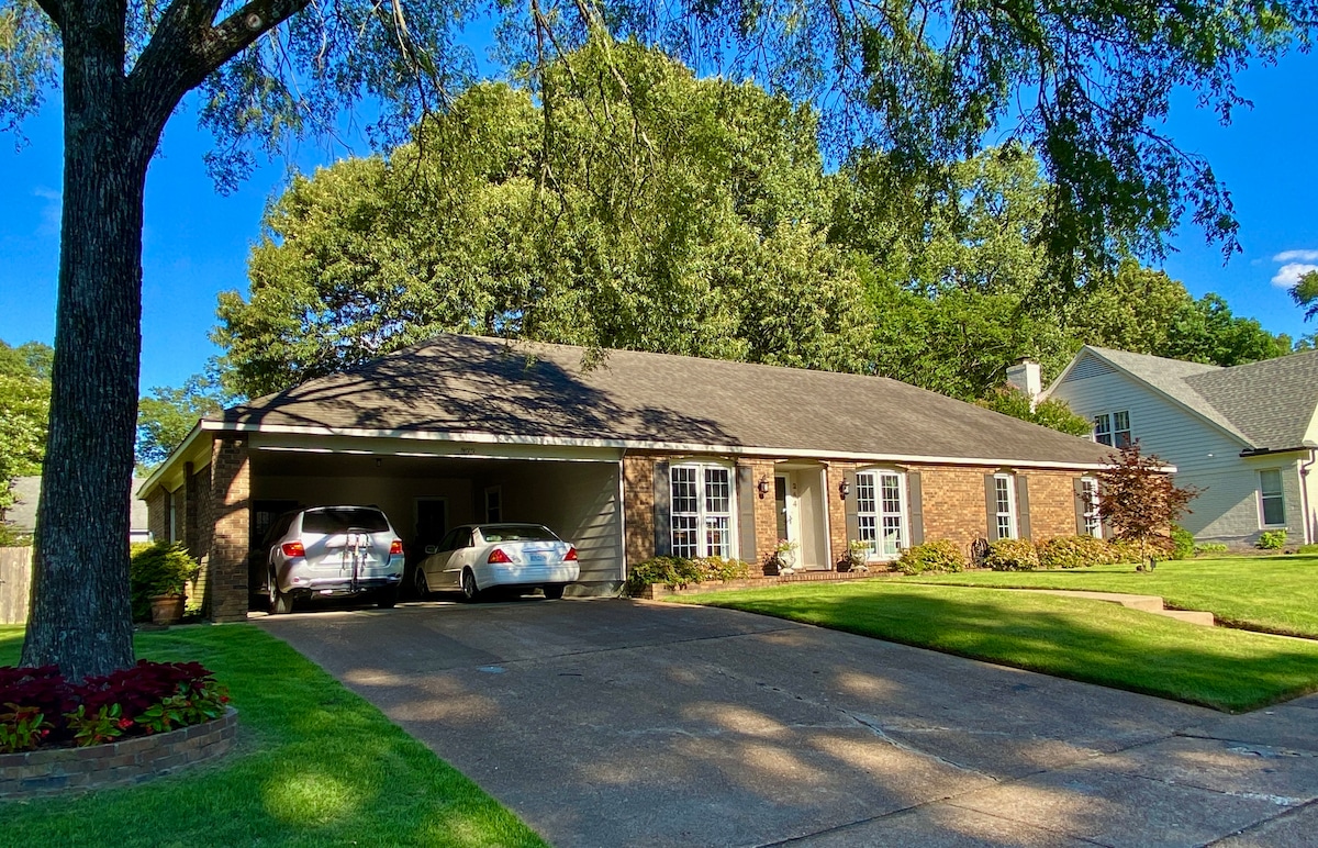 The exterior of a single-story brick home is presented, featuring a spacious driveway with two parked cars. Lush green grass surrounds the home, and a large tree provides shade to the front yard. The house is positioned within a quiet, tree-lined neighborhood.