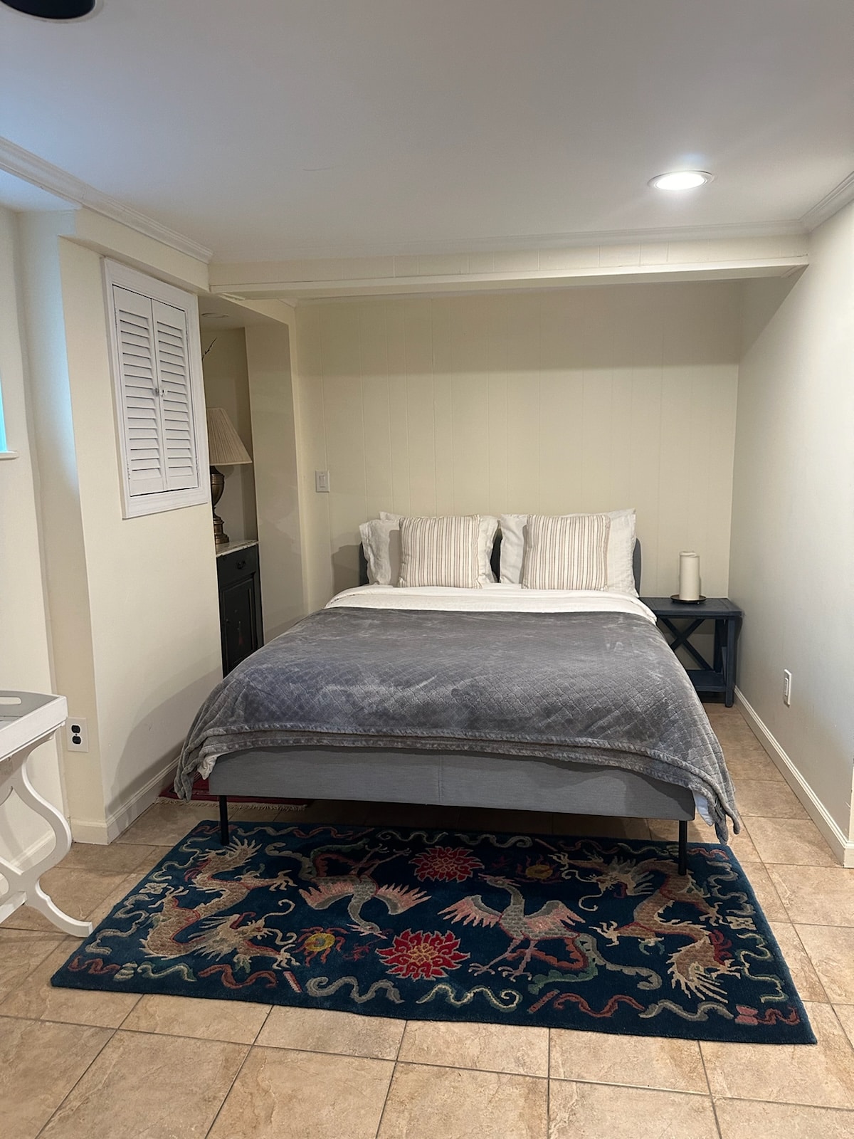 A cozy sleeping area features a spacious bed with soft linens and decorative pillows. A textured gray blanket is draped across the bed. A colorful area rug occupies the floor, while natural light enters through a window. A small side table is positioned nearby.