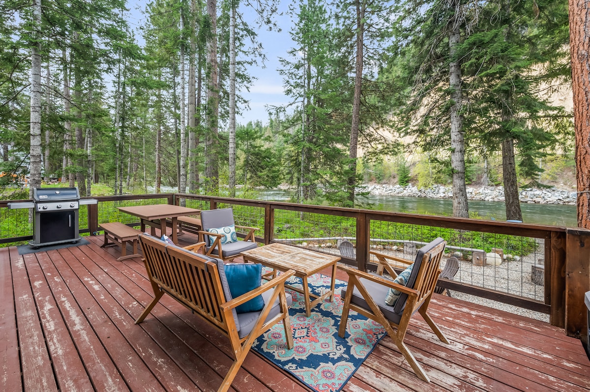 A spacious deck extends over natural wooden planks, featuring outdoor seating arrangements including a table and lounge chairs. A grill is positioned nearby, and the surrounding greenery offers views of trees and a river, creating a serene outdoor environment.