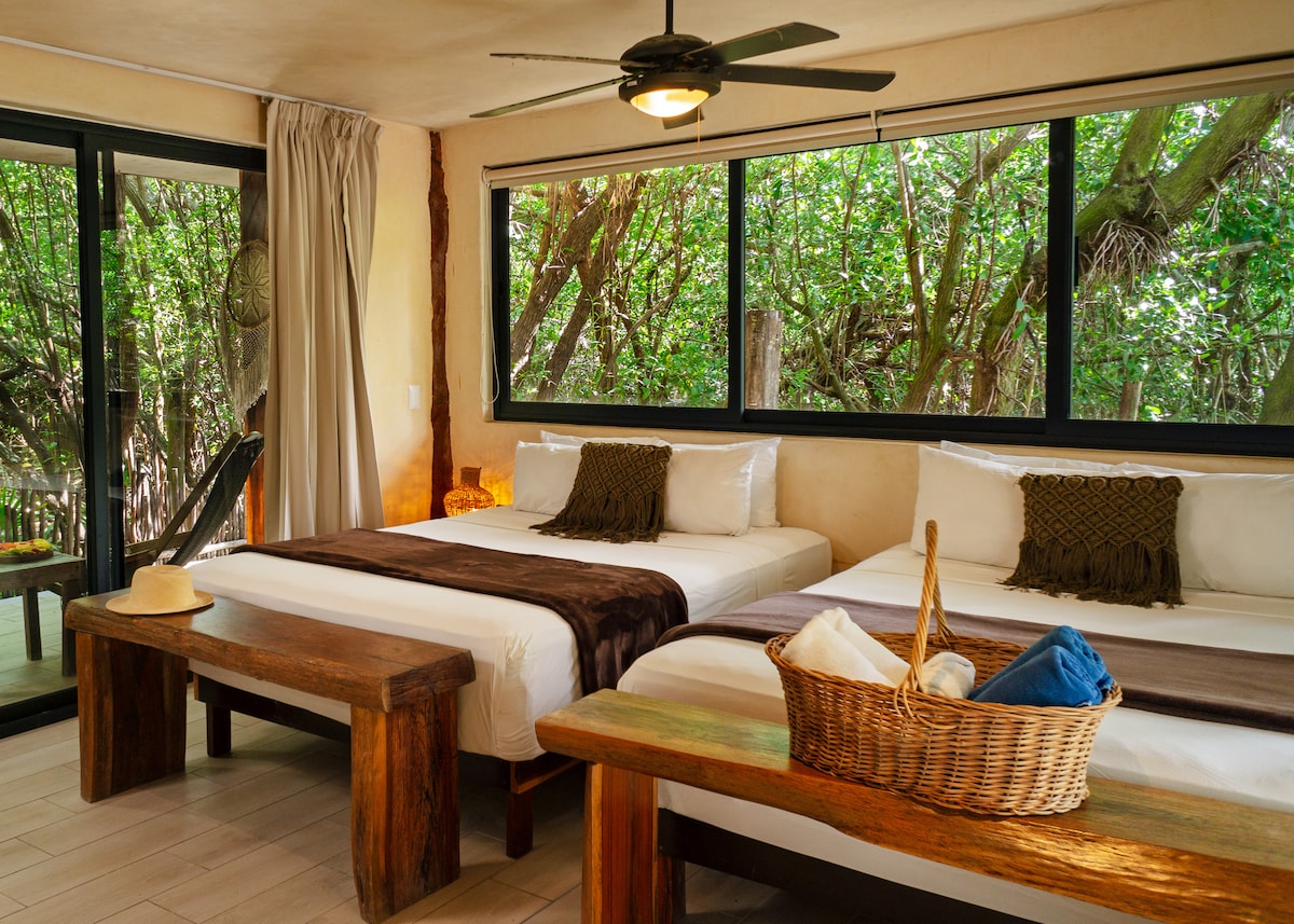 A spacious room features two queen size beds dressed in organic cotton linens. Large windows reveal views of lush greenery, while a wooden table and chairs offer a place for relaxation. Natural light fills the room, complemented by soft decor elements and a ceiling fan.