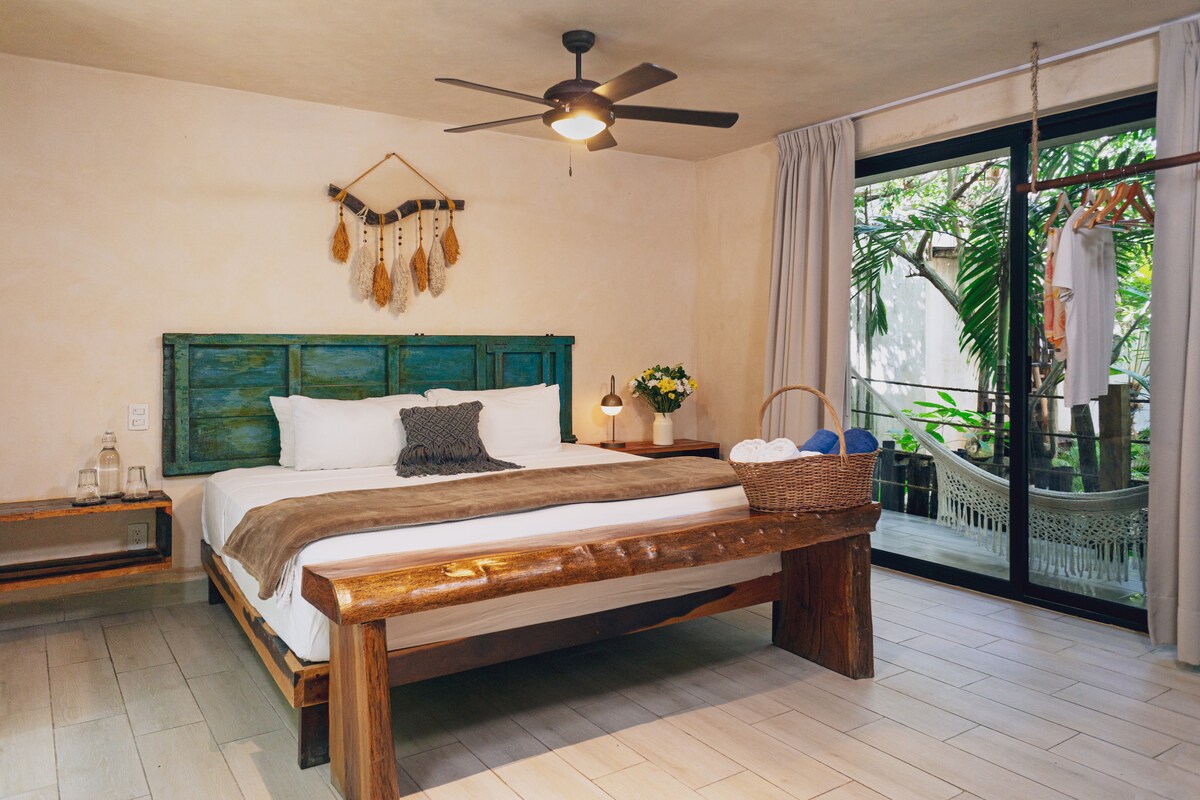 A spacious room features a king-size bed with a light brown throw blanket and decorative pillows. Natural light enters through large windows, revealing a private balcony with a hammock. Wooden furniture includes a bedside table and a stylish wall hanging provides a touch of texture.