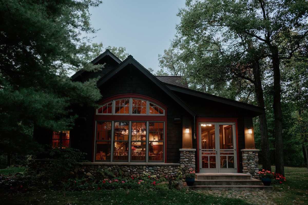 A charming cabin exterior is framed by lush greenery, showcasing large windows that illuminate a warm interior. Stone accents and soft outdoor lighting enhance the inviting entrance, which features double doors and a colorful display of potted flowers along the pathway.