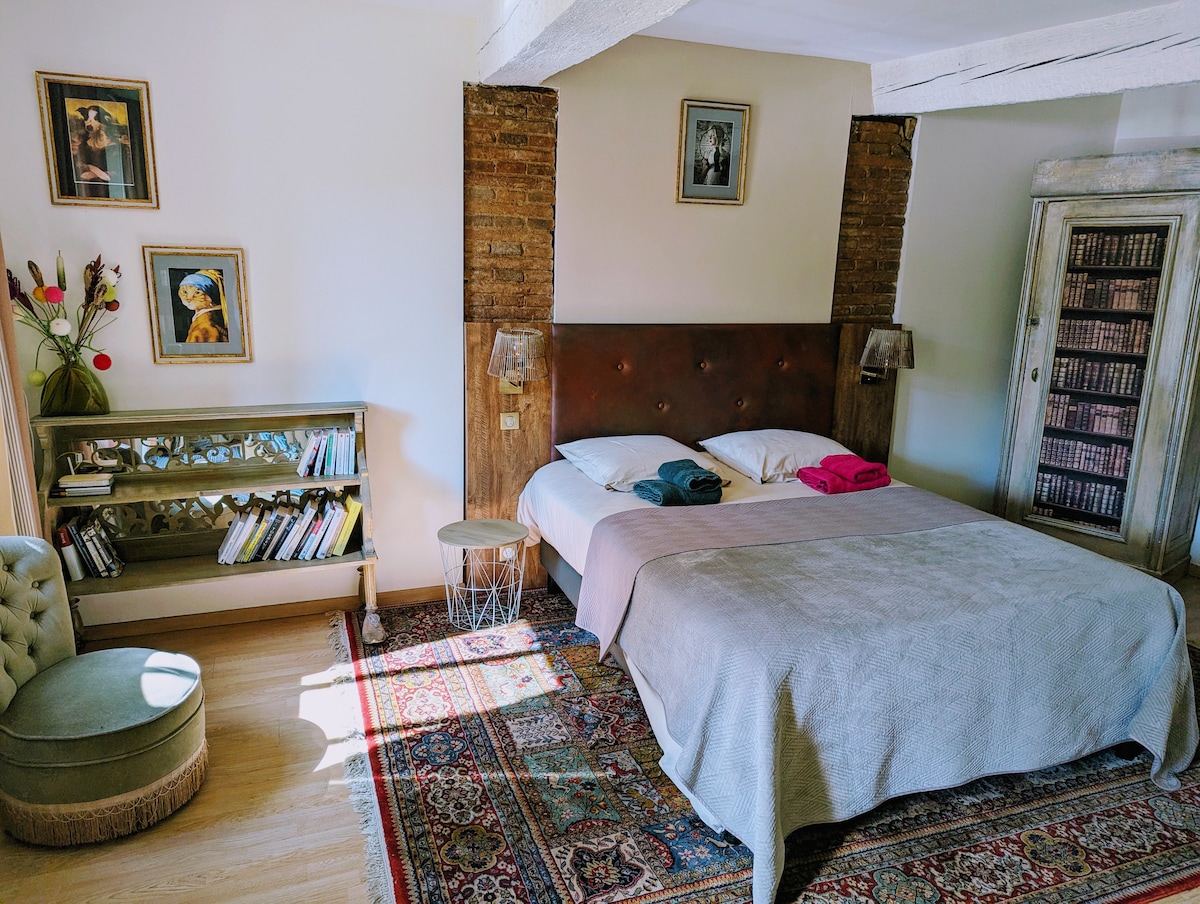 A spacious bedroom is presented with a large bed, dressed in neutral linens and accompanied by fluffy pillows. Decorative elements include framed art on the walls and a stylish bookshelf. A comfortable chair sits beside a small side table, enhancing the inviting ambiance.