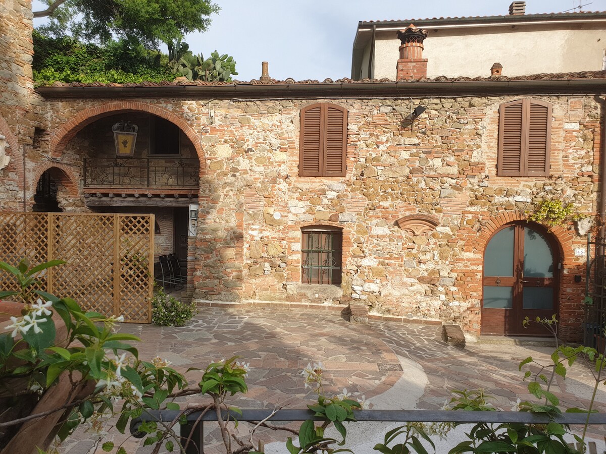 An old stone building, featuring a rustic façade with wooden shutters, stands in a courtyard. A decorative archway and a wooden door are visible, surrounded by greenery. The courtyard is paved with stone, blending harmoniously with the historical character of the property.
