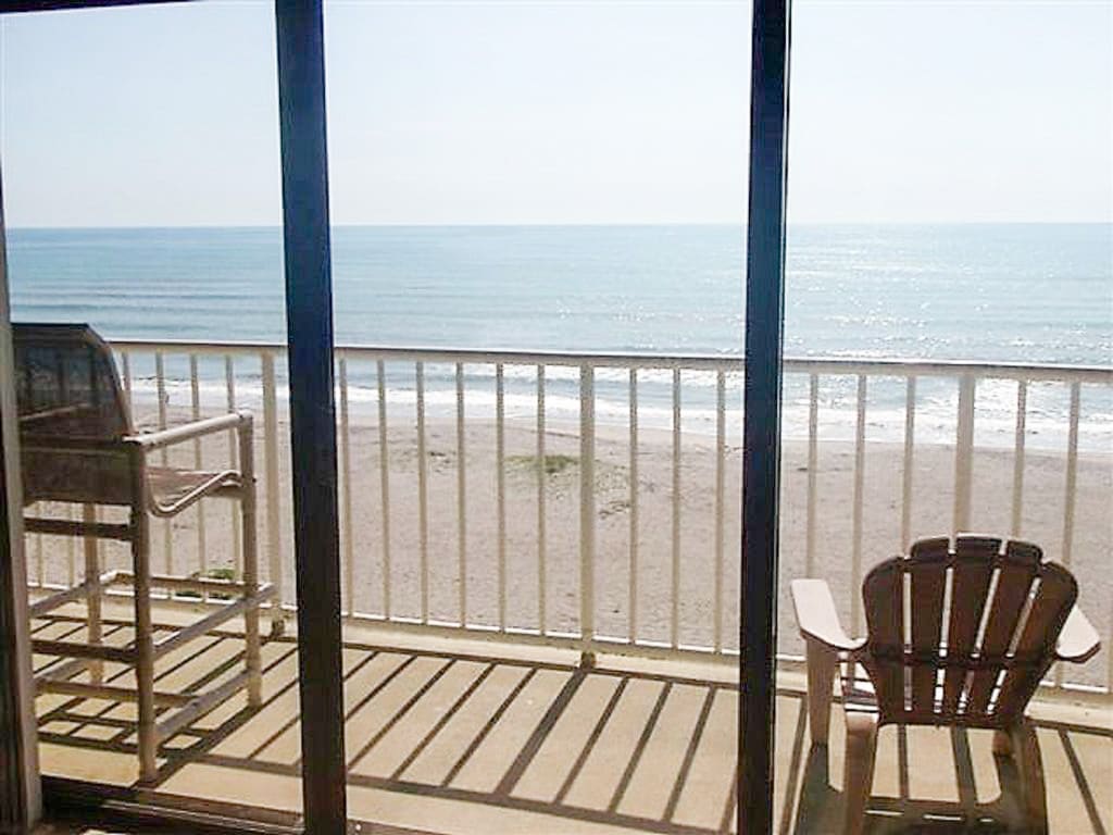 A wide balcony with a view of the sandy beach and ocean is showcased. Two outdoor chairs are positioned, with sunlight casting shadow patterns on the floor. The ocean's gentle waves can be seen extending to the horizon under a clear sky.