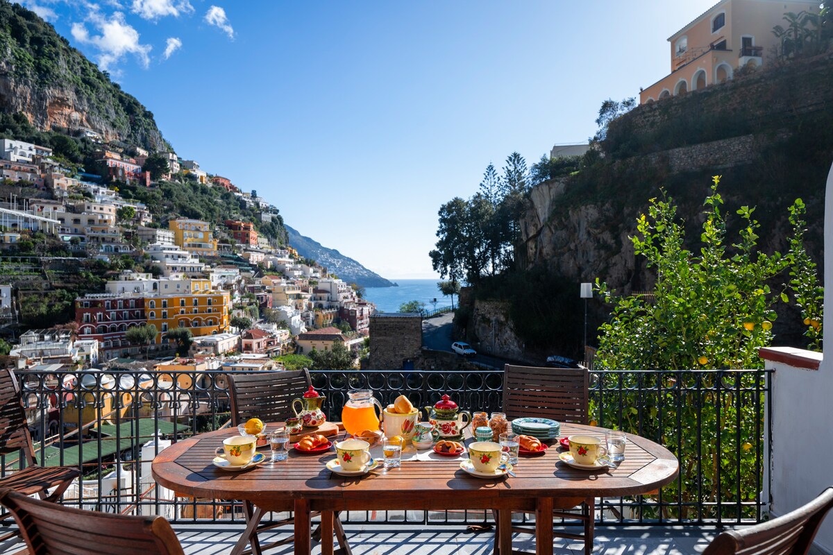 A terrace table is set for breakfast, featuring colorful dishes and a refreshing beverage. The backdrop reveals the scenic Positano coastline, with vibrant buildings climbing the hillside, and glimpses of the sea visible through the greenery.