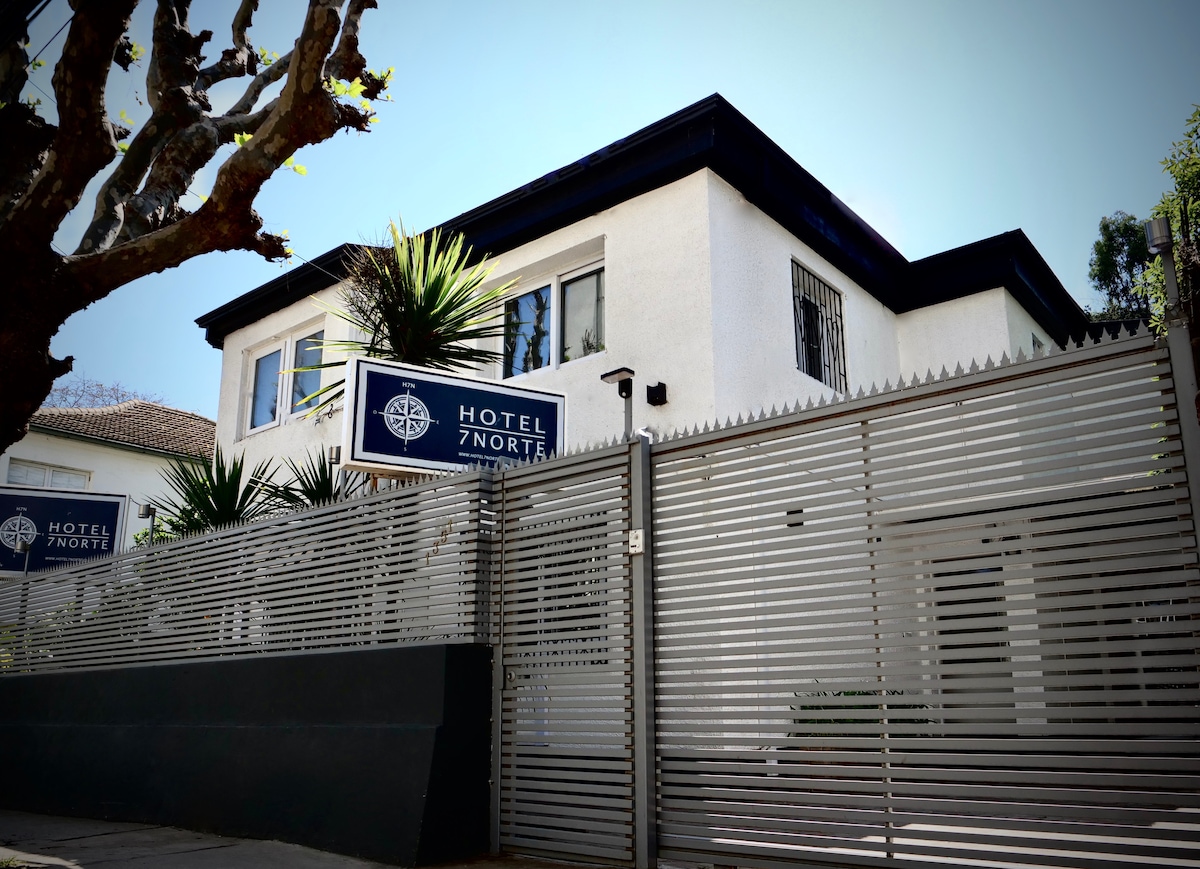 A modern, white building with a flat roof and large windows is surrounded by greenery. A gated entrance with horizontal metal slats provides a sense of privacy. The hotel's sign is prominently displayed, indicating its name.