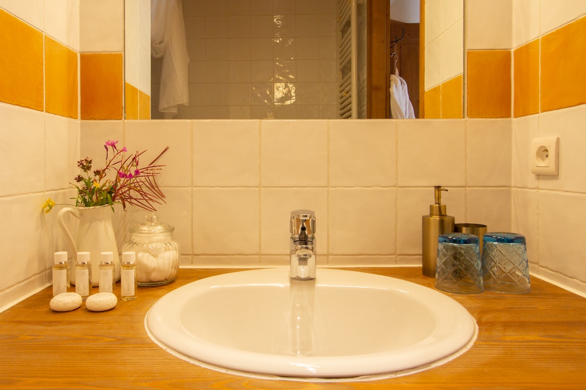 A clean and functional bathroom sink area is displayed, featuring a circular basin set on a wooden countertop. Touches of décor, including a vase of flowers and glass containers, enhance the space. The mirror reflects the light, contributing to the overall bright ambiance.