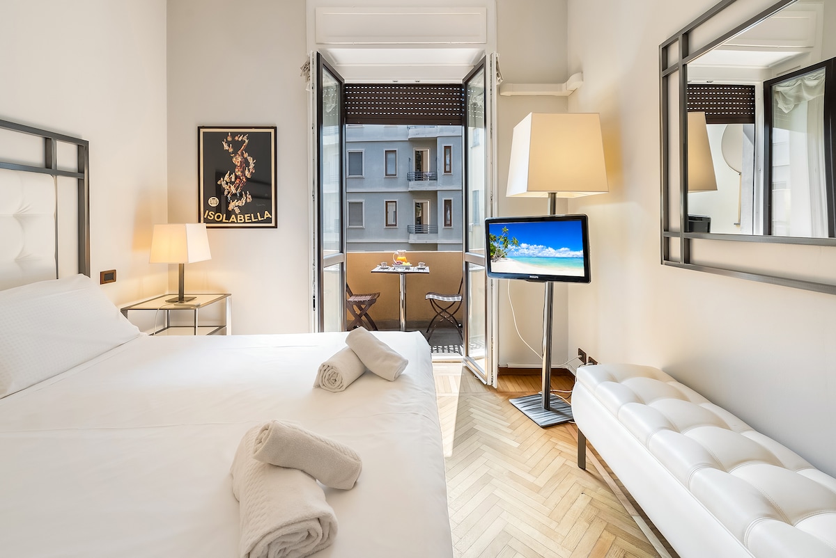 A bright bedroom features a spacious bed with neatly arranged towels. Open doors lead to a small balcony with two chairs and a table. A television and a floor lamp are positioned adjacent to the bed, with a large mirror reflecting the room's elegant decor.