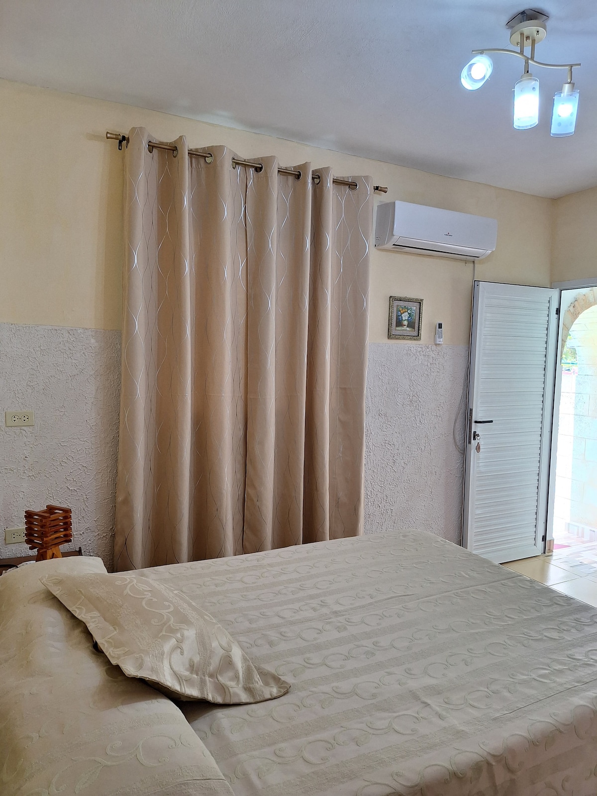 The room features a spacious bed with a light-colored coverlet. Soft beige curtains hang beside a door leading to the outside. A wall-mounted air conditioning unit and a framed picture are visible, contributing to the room's comfort.