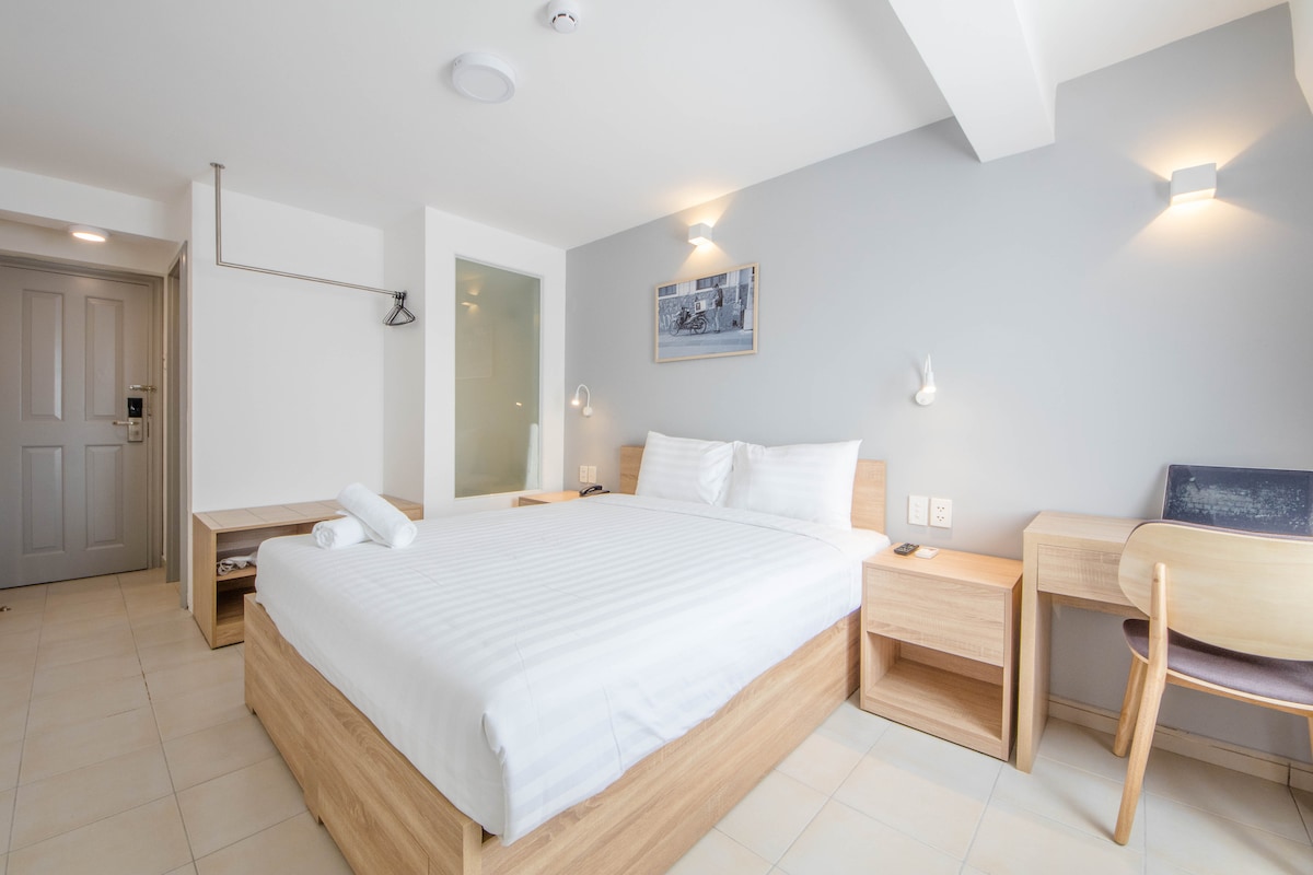 A clean and bright room features a spacious double bed with crisp white linens. Two wooden bedside tables are positioned on either side. A work desk and chair are present, along with soft lighting on the walls. A glass partition leads to the private bathroom.