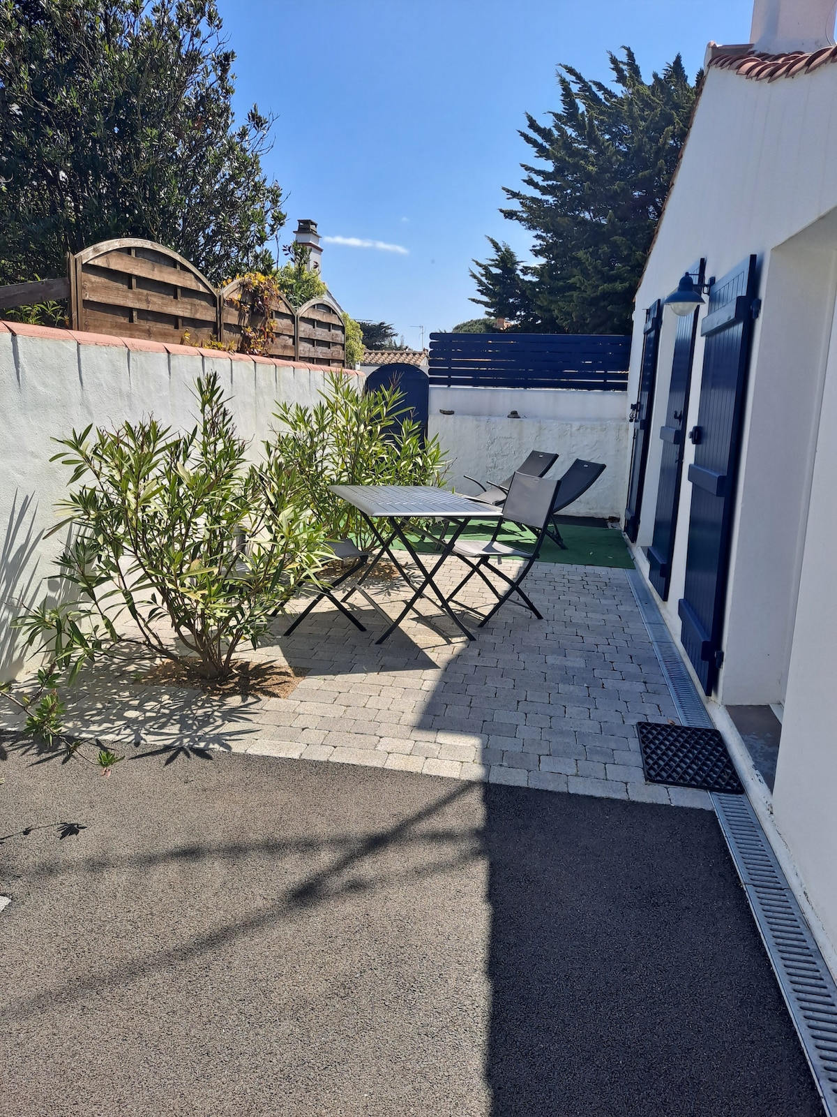 A private terrace area is shown, featuring a small table set with two foldable chairs. Lush greenery, including a young olive tree, borders the space, and fencing provides privacy. The ground is paved with light-colored stones and dark rubber matting.