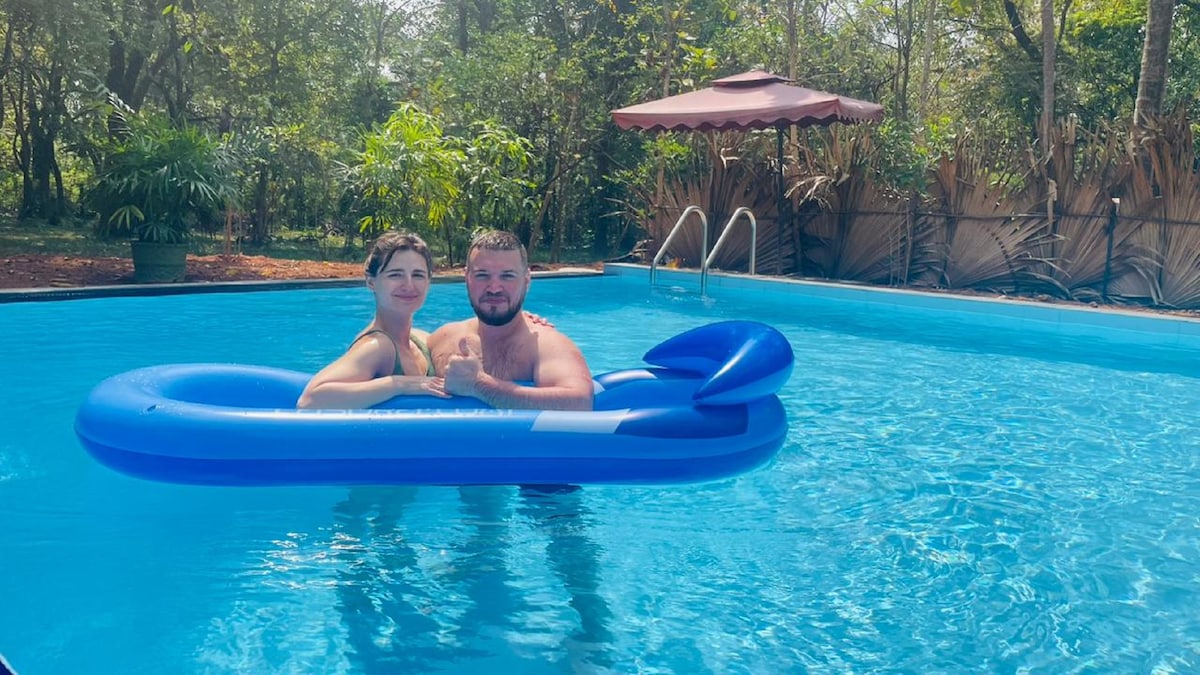 A large swimming pool is framed by lush greenery, offering a serene outdoor space. Two individuals are seated on a blue float, enjoying the clear water. A shaded area with an umbrella is visible along the poolside, enhancing relaxation options.