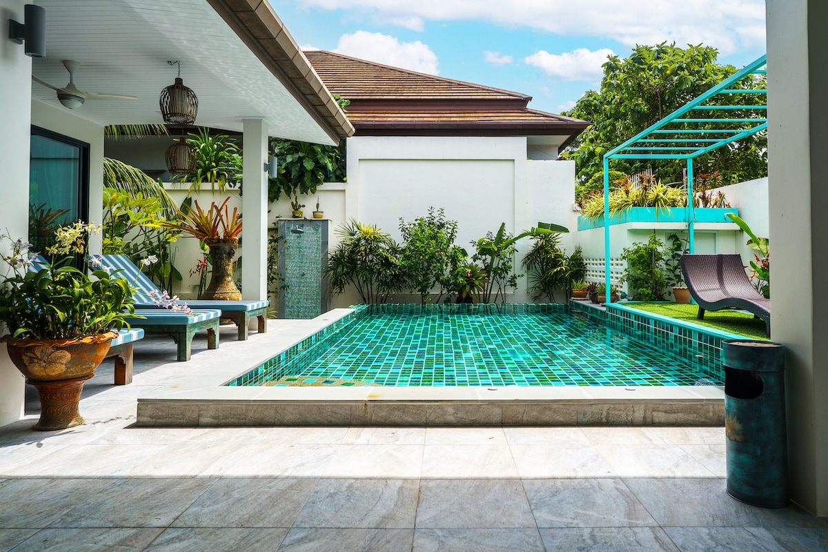 A private swimming pool is surrounded by vibrant tropical plants and decorative pots. Sun loungers placed on a tiled terrace invite relaxation. A shaded area features a pergola, complementing the contemporary design of the villa. Natural light highlights the bright turquoise tiles of the pool.