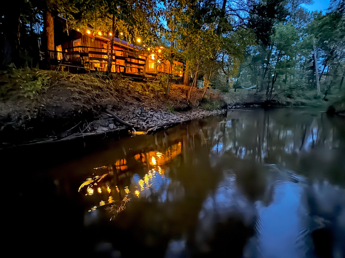 The cabin is softly illuminated by warm lights, reflecting gently on the calm surface of the river. Surrounding trees create a natural framing, enhancing the private and tranquil atmosphere of the setting as dusk settles in.