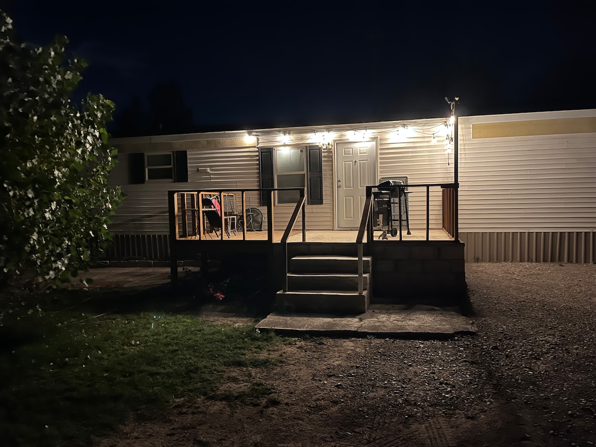 A mobile home exterior is illuminated by string lights, highlighting a welcoming porch with a set of chairs and a grill. Light spills from a central door, creating an inviting entryway. The ground is gravel, with a grassy area visible in front.
